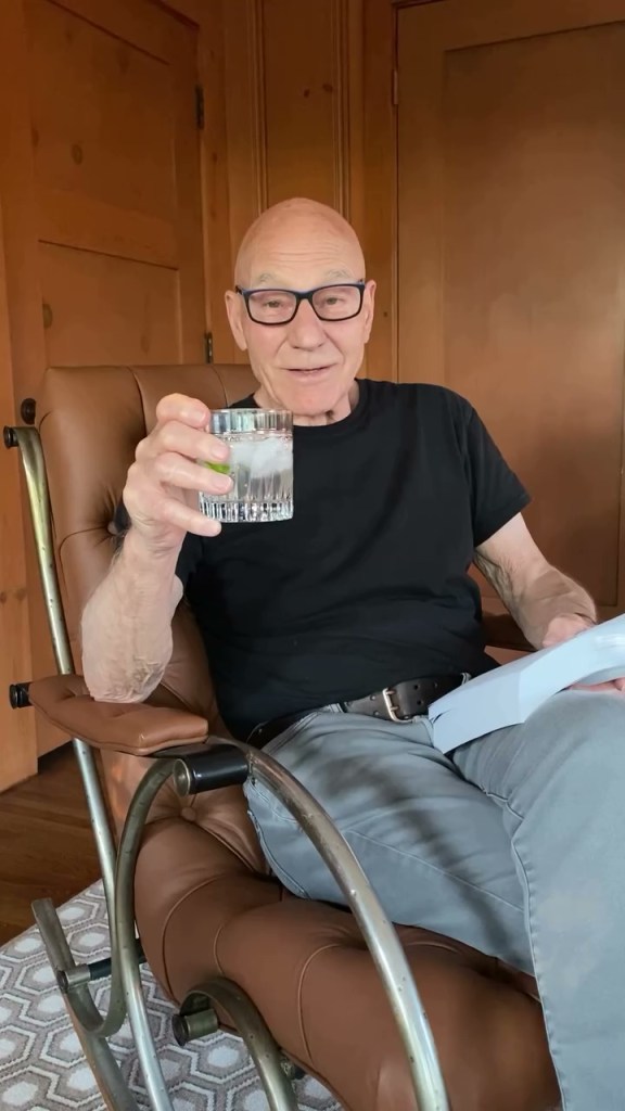 Patrick Stewart sitting in a chair, holding a drink and a book.
