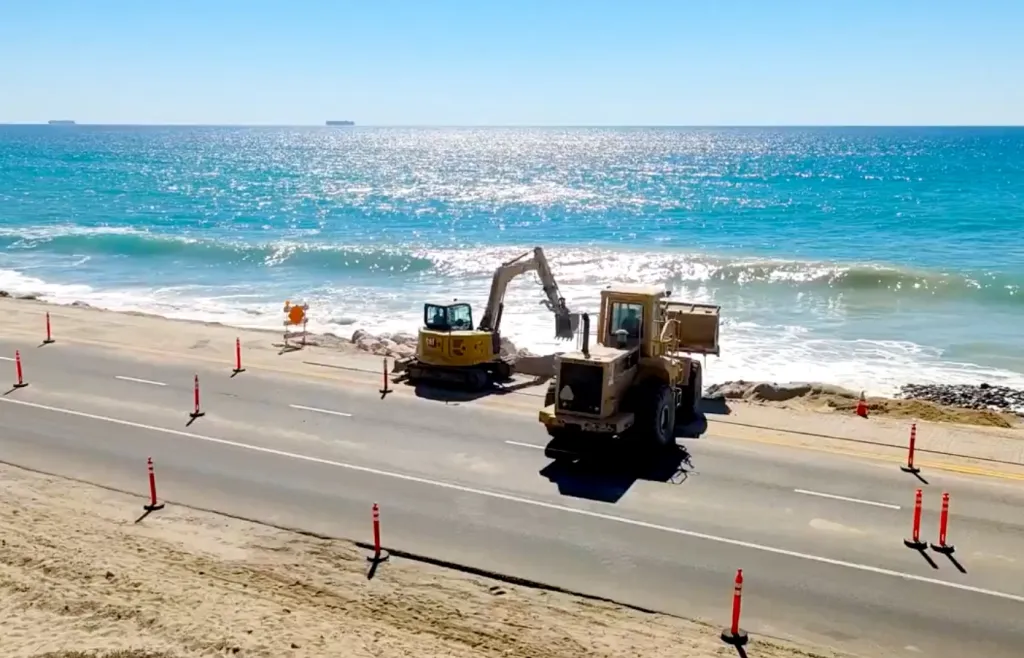 Construction equipment building a rock slope along a coastal highway next to the ocean.