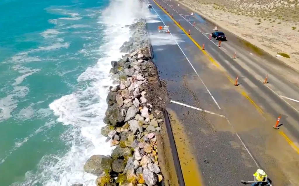 Aerial view of crews constructing Rock Slope Protection along State Route 1 at Thornhill Broome Beach, where waves crash against the newly placed rocks next to the highway.