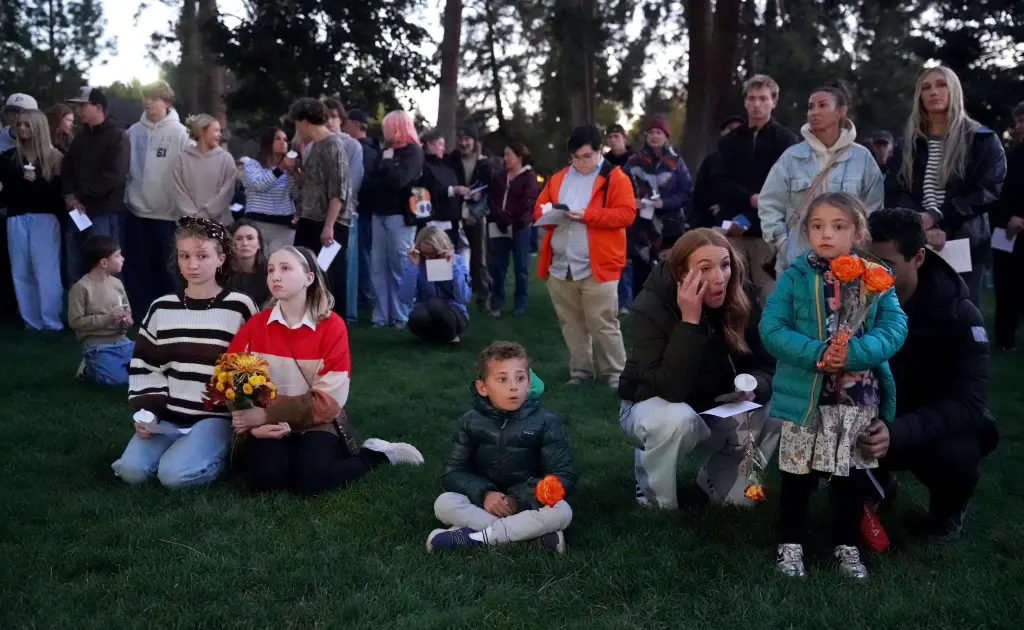 People gather in a park for a candlelight vigil, with some holding flowers and candles while seated and standing on the grass.