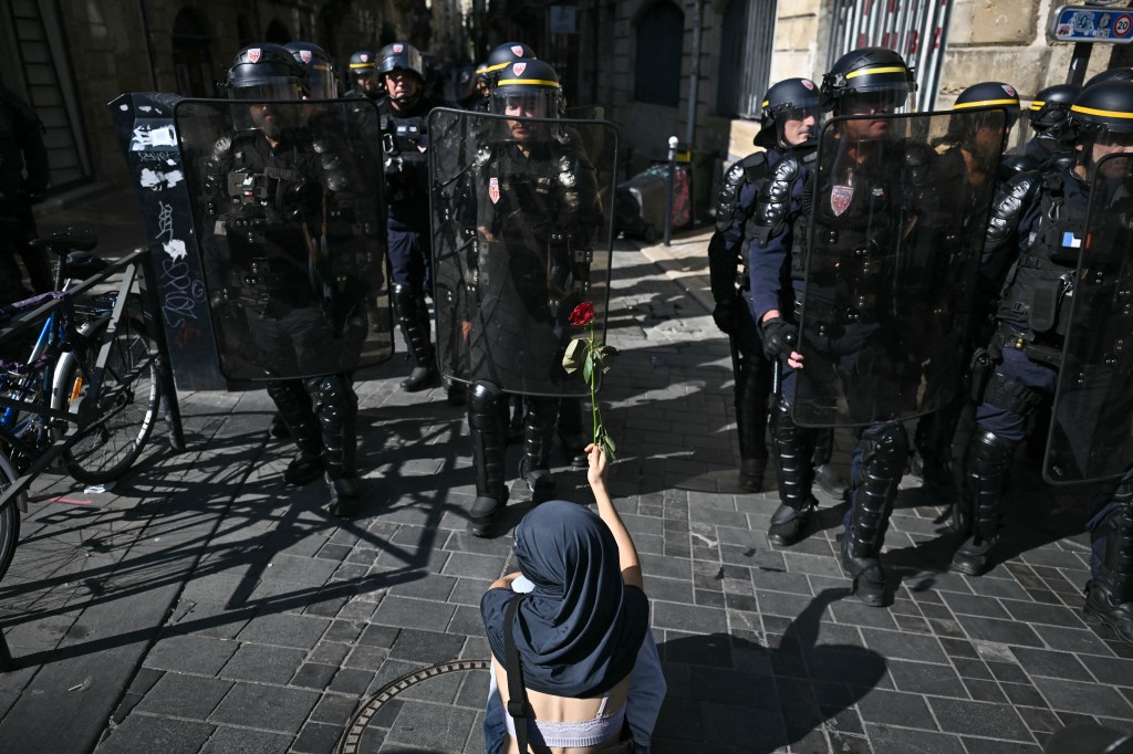 A person holding a red rose faces a line of riot police in full gear.