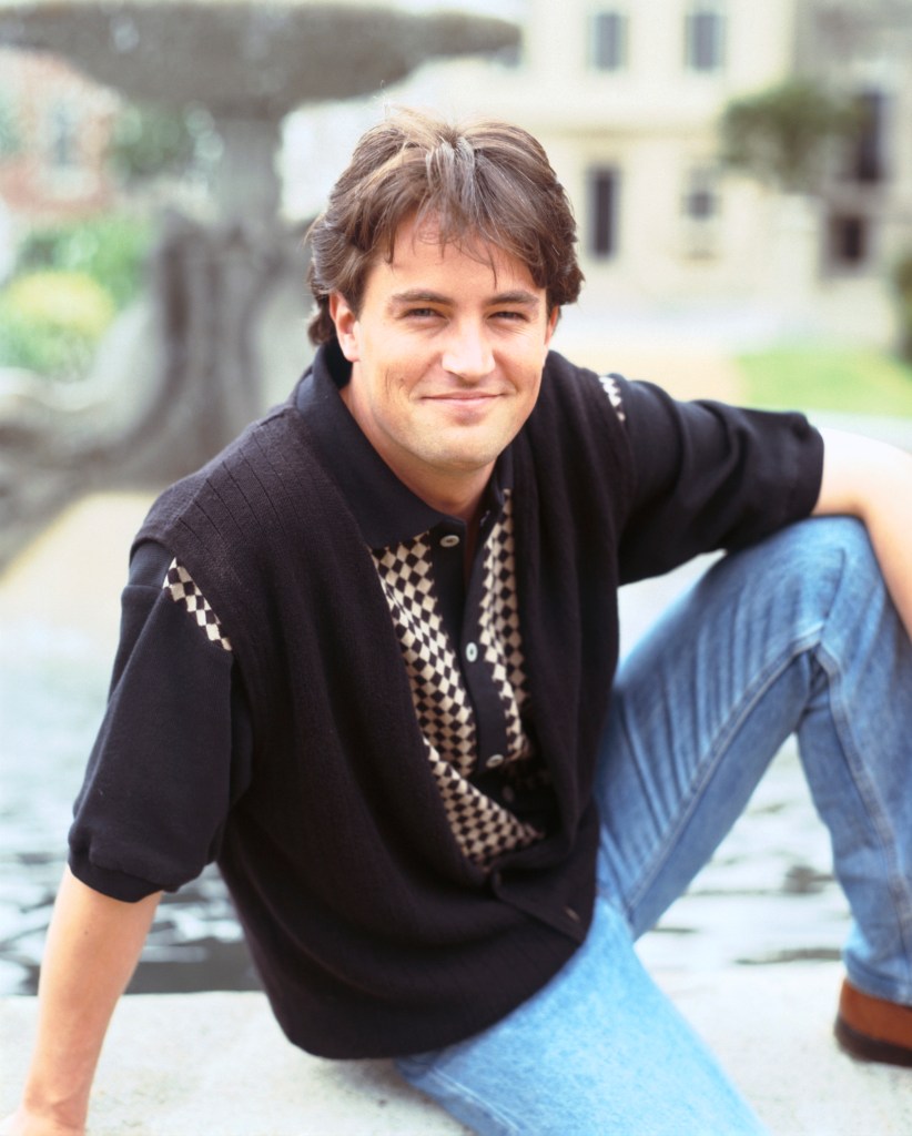 Matthew Perry as Chandler Bing smiling outdoors, wearing a black shirt and jeans.