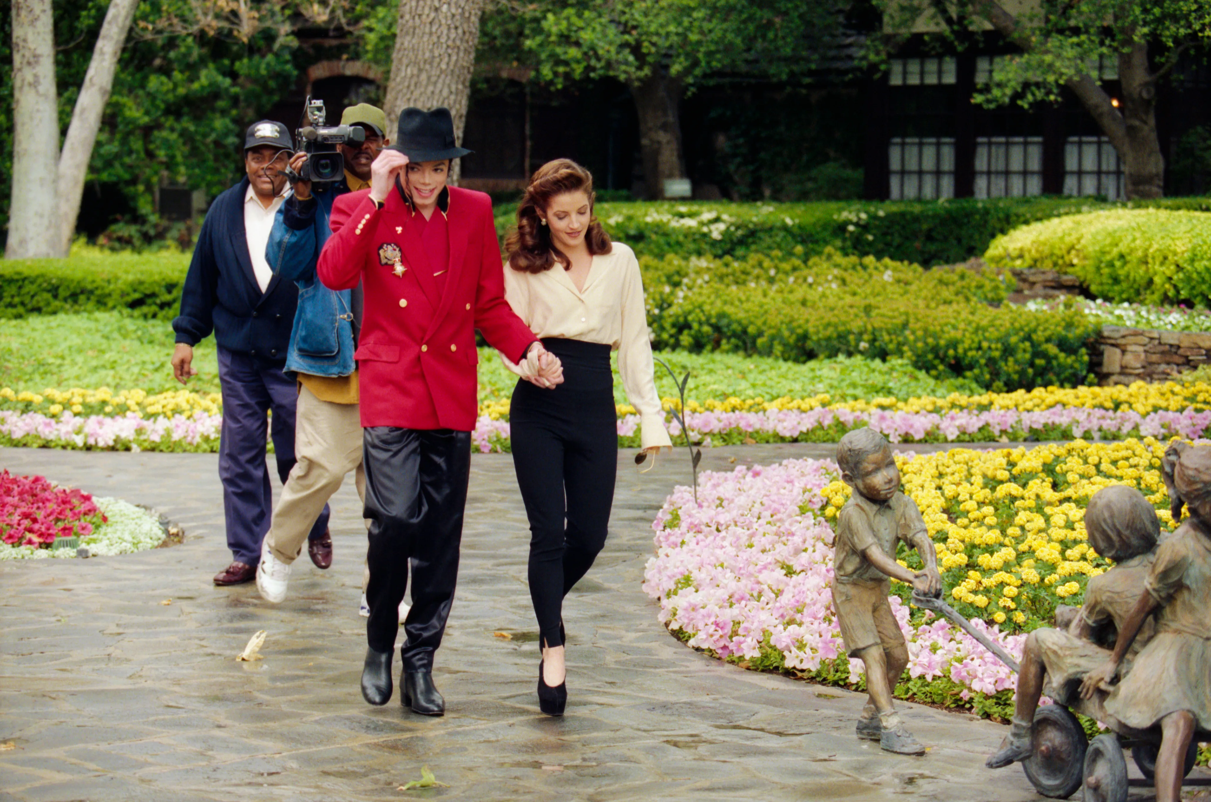 Michael Jackson and Lisa Marie Presley holding hands while walking on a stone path at Neverland Ranch, with a camera operator and another person following behind them.
