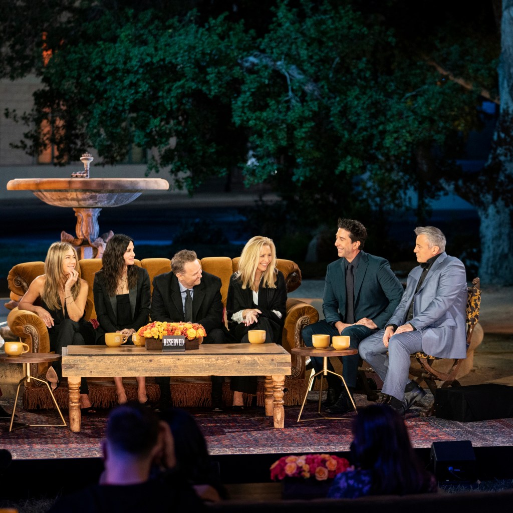 The cast of Friends seated on a couch in front of the iconic fountain outdoors at night.