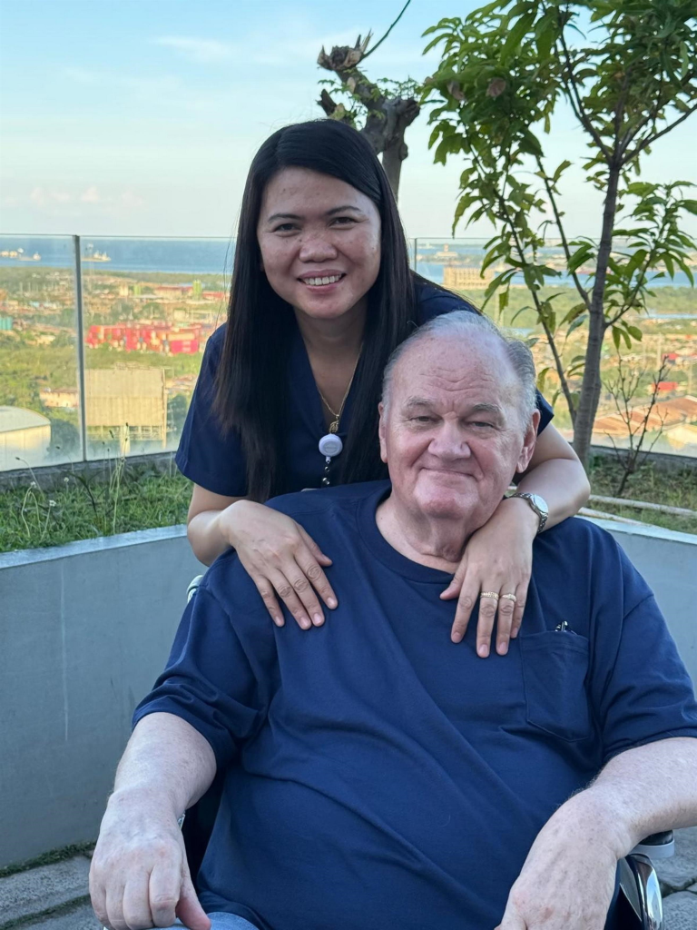 Thomas Markle smiling while seated in a wheelchair outdoors with a woman standing behind him, her hands resting on his shoulders.