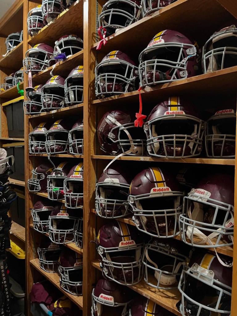 Wooden shelves filled with maroon and gold football helmets with grey facemasks.