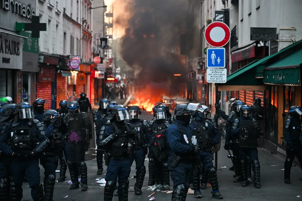 Police officers in riot gear stand in a Paris street in front of a fire and thick smoke.
