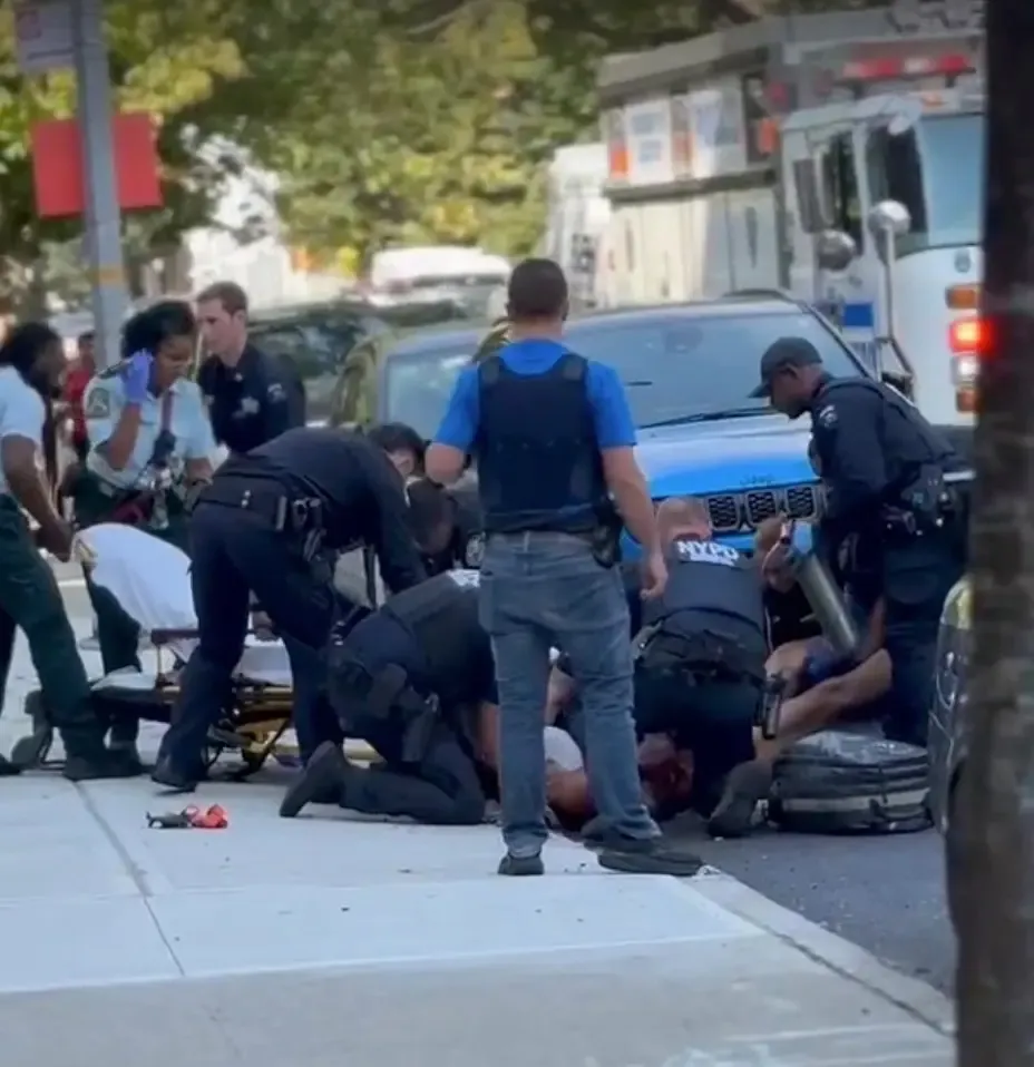Police officers surround a wounded colleague near a blue vehicle on a city street.