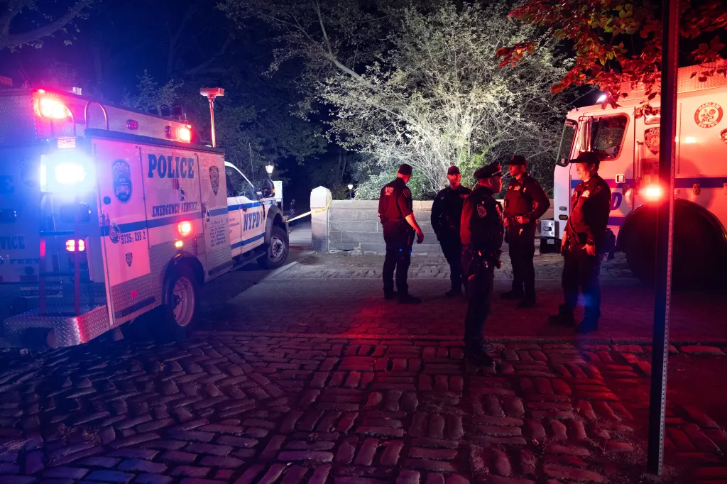 Police officers at a crime scene at night, flanked by emergency vehicles.