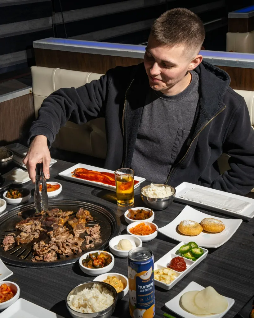 A customer uses tongs to grill meat at a table filled with various side dishes and drinks at a Gen Korean BBQ restaurant.