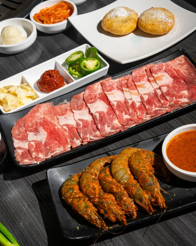 A spread of Korean BBQ dishes including thinly sliced raw beef, seasoned shrimp, side dishes of kimchi and garlic, and two pastries dusted with powdered sugar.