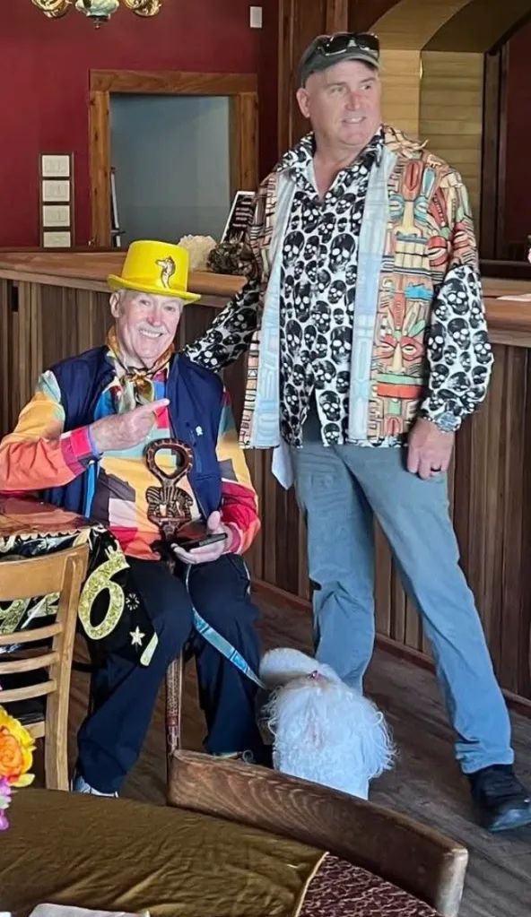 Tom O'Bryan stands next to a seated man wearing a yellow hat and colorful clothing, with a small white dog nearby.