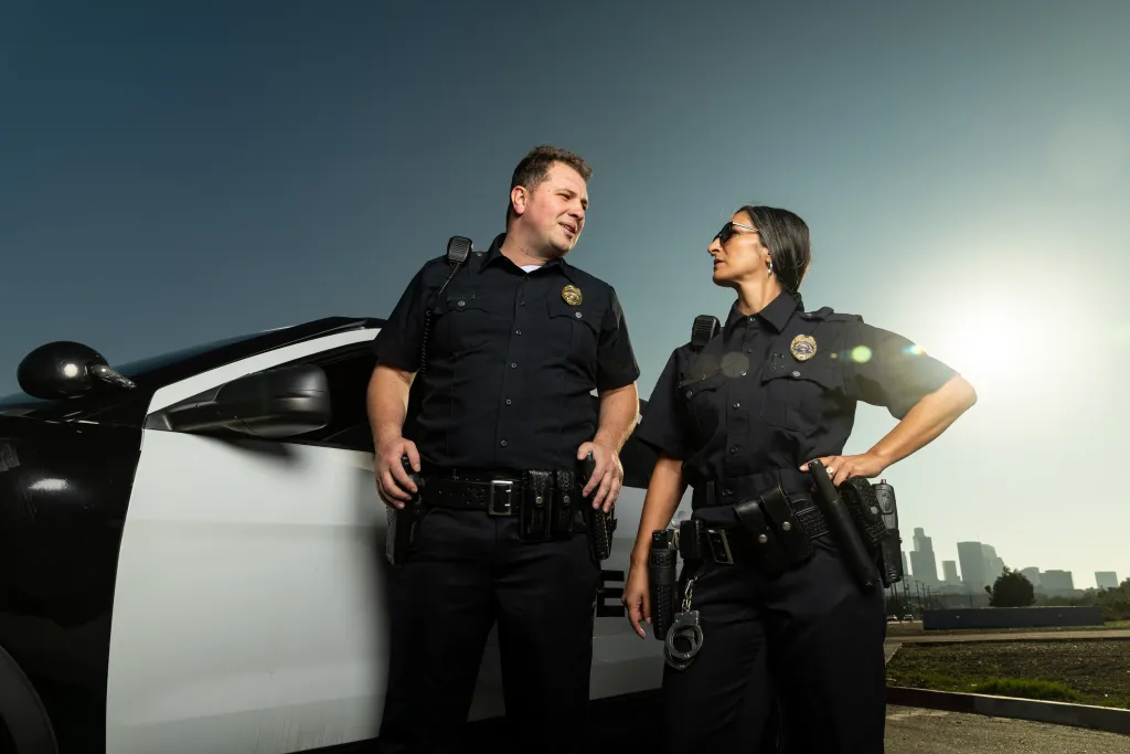 A Southeast Asian female police officer and a white male police officer talk while standing next to a police squad car.