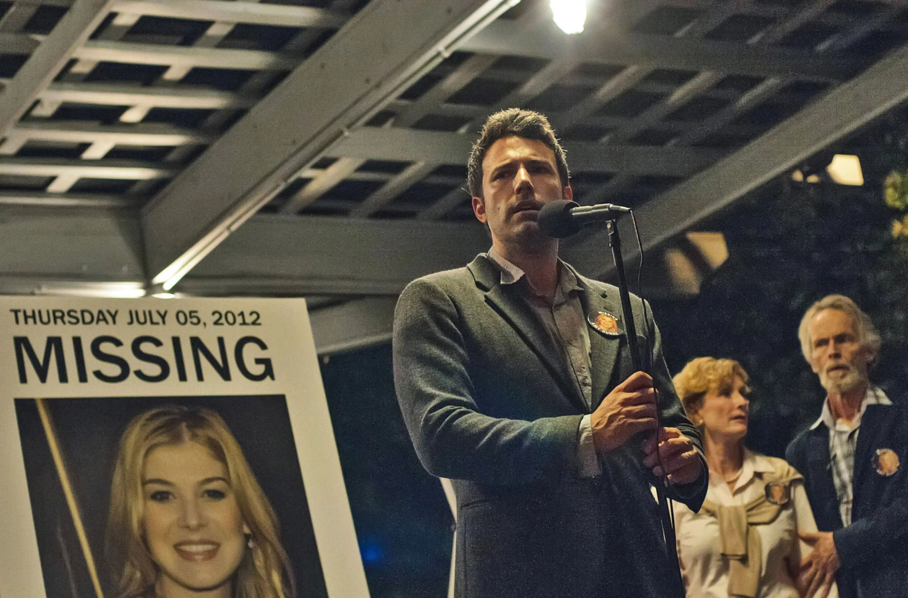 Ben Affleck speaks into a microphone at a podium next to a missing person poster of Rosamund Pike, with Lisa Banes and David Clennon standing behind him.