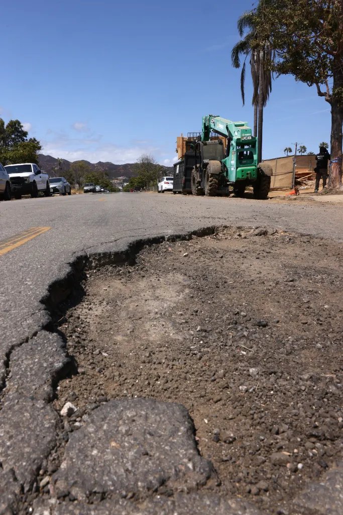 A large, deep pothole in the asphalt of a street, with a green telehandler and construction vehicles parked in the background.