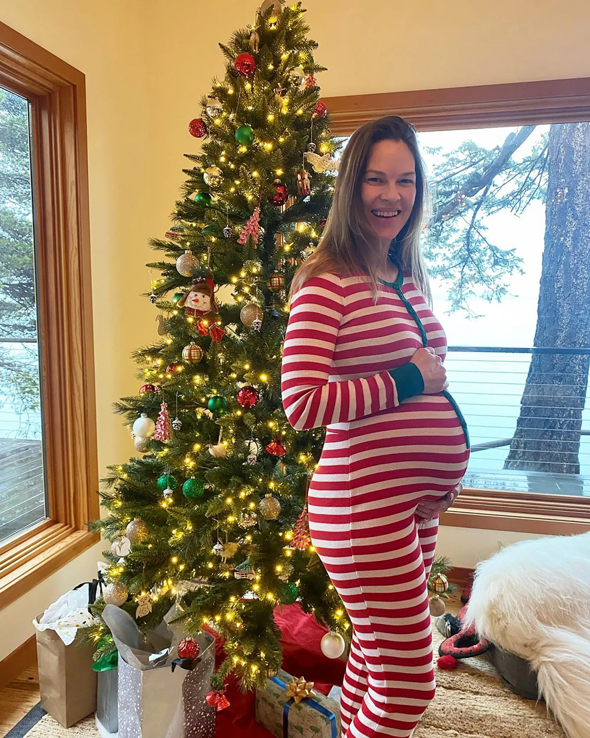 Hilary Swank stands smiling in front of a decorated Christmas tree, wearing red and white striped pajamas and cradling her pregnant belly.