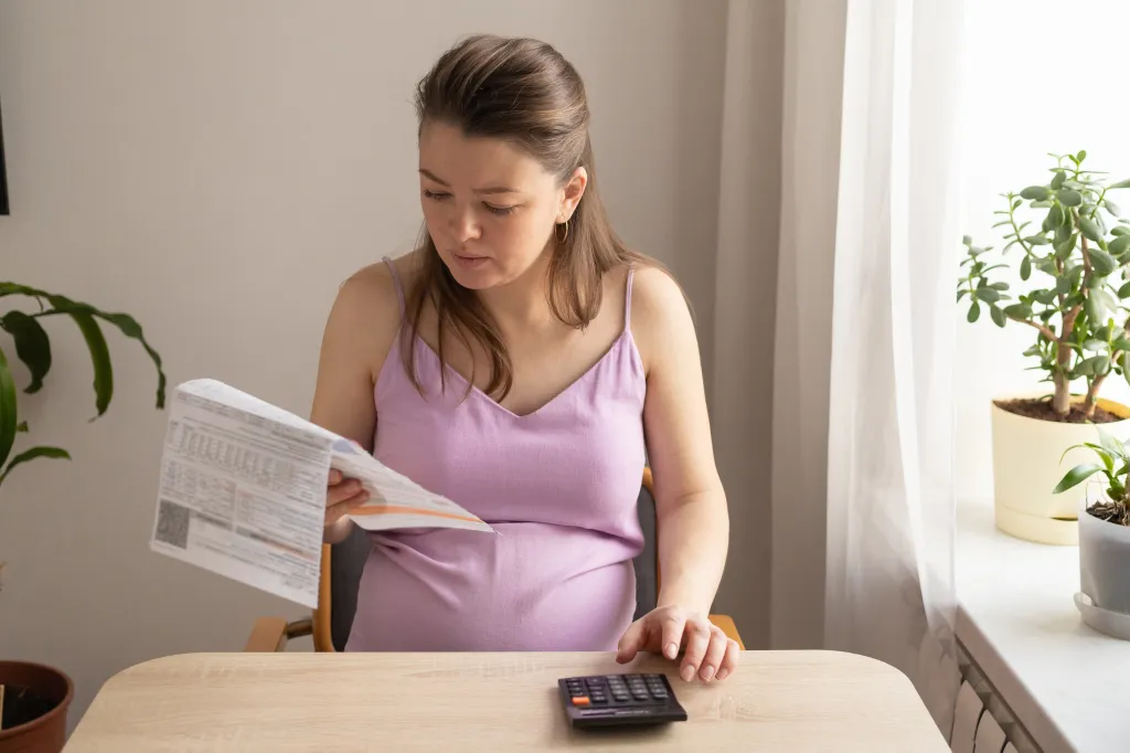 A pregnant woman sits at a table, holding a utility bill while using a calculator to manage household finances.