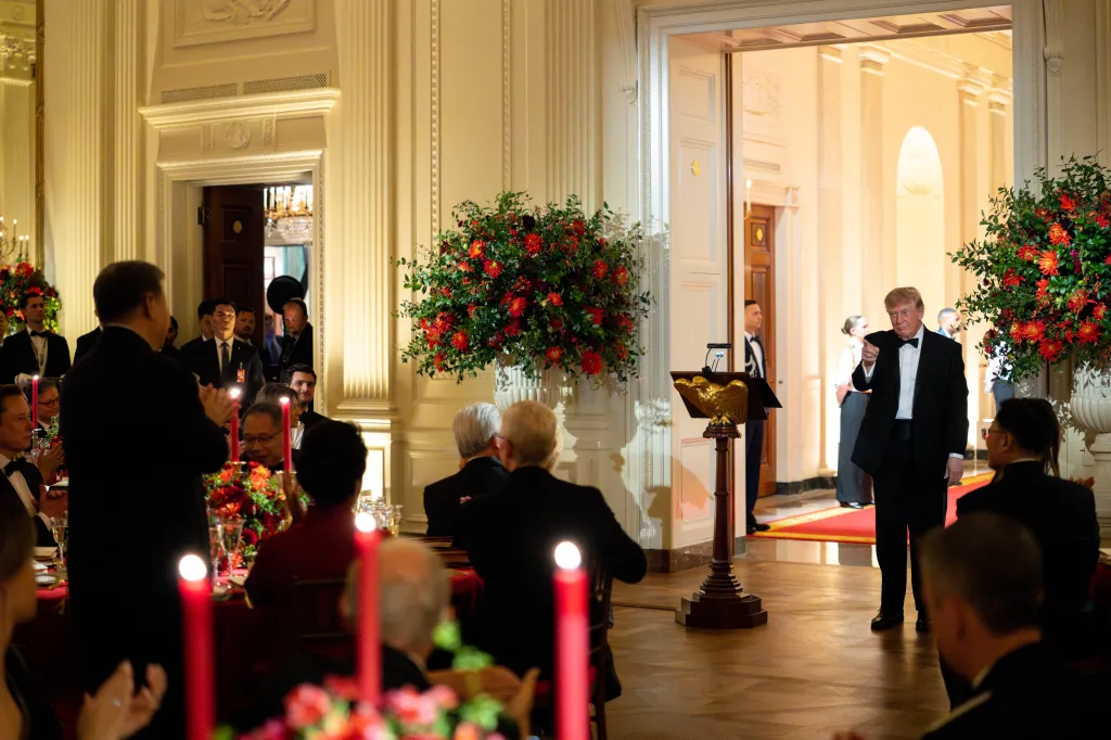 President Donald J. Trump stands in a doorway, gesturing to guests seated at tables adorned with red flowers and lit candles during a State Dinner in the East Room of the White House.