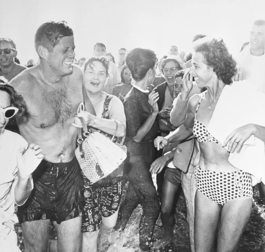 President John F. Kennedy, shirtless in swim trunks, is surrounded by an excited crowd of admirers in the water at Santa Monica, California.