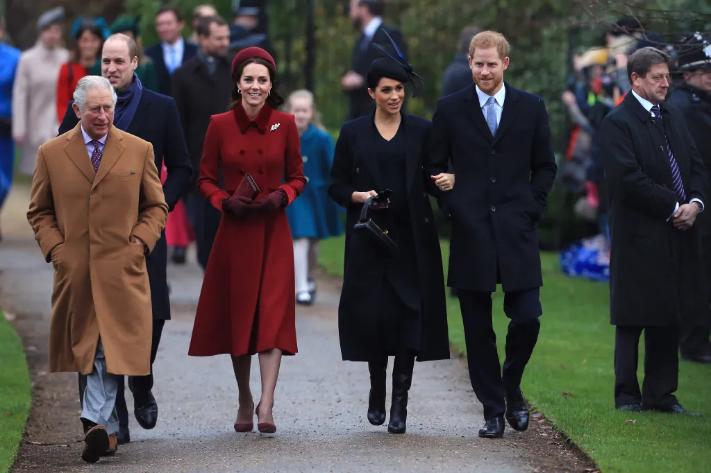 Prince Charles, in a brown oercoat, Prince William, in a black coat, Kate Middleton in a red coat and red hat, Meghan, Duchess of Sussex, in a black coat and black hat, and Prince Harry, in a black coat, walking together outdoors.