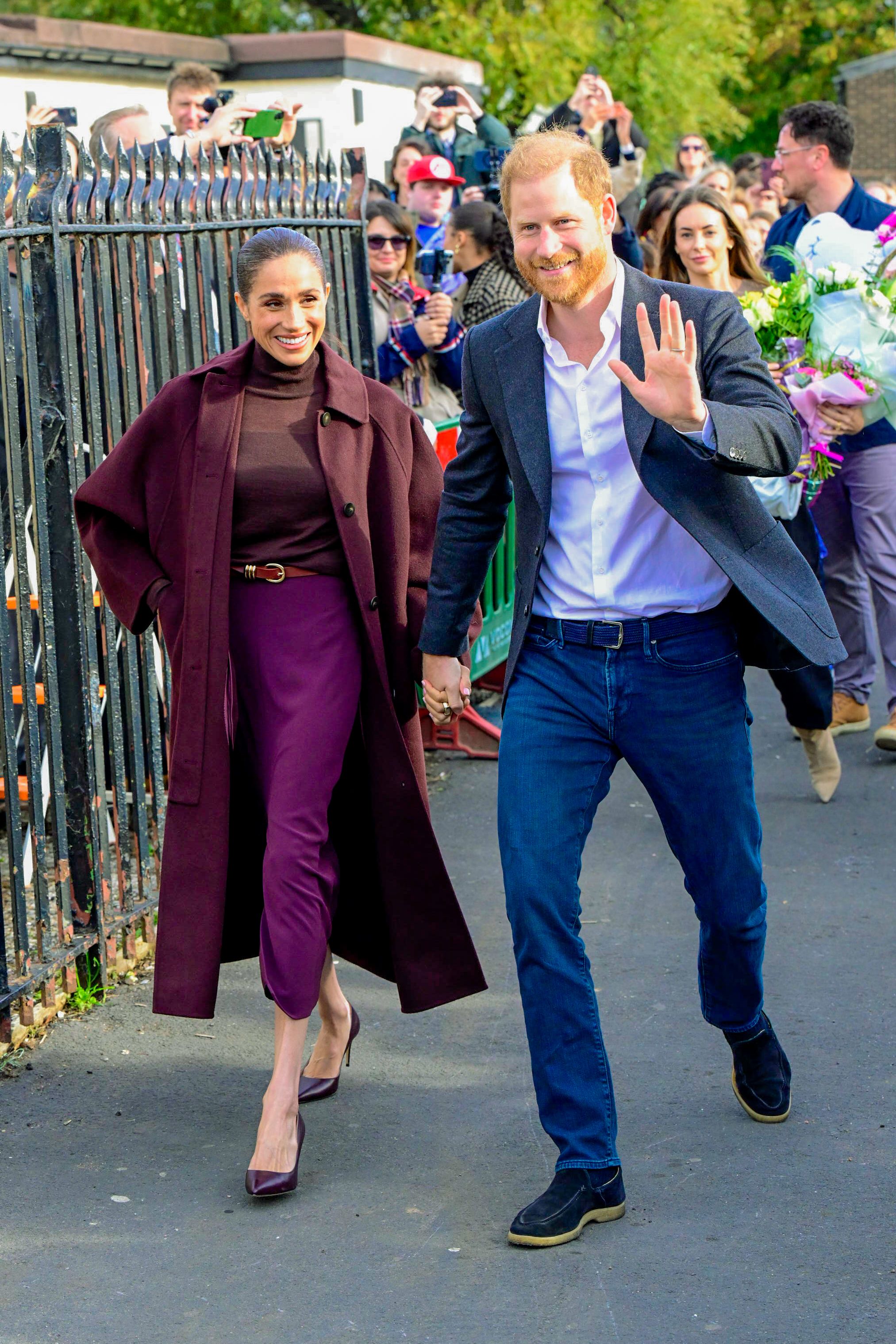 Prince Harry and Meghan Markle walking hand in hand, with Prince Harry waving to a crowd in the background.
