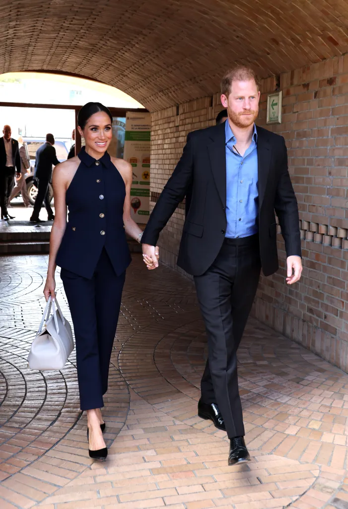 Prince Harry, Duke of Sussex, and Meghan, Duchess of Sussex, walk hand in hand through a brick archway.