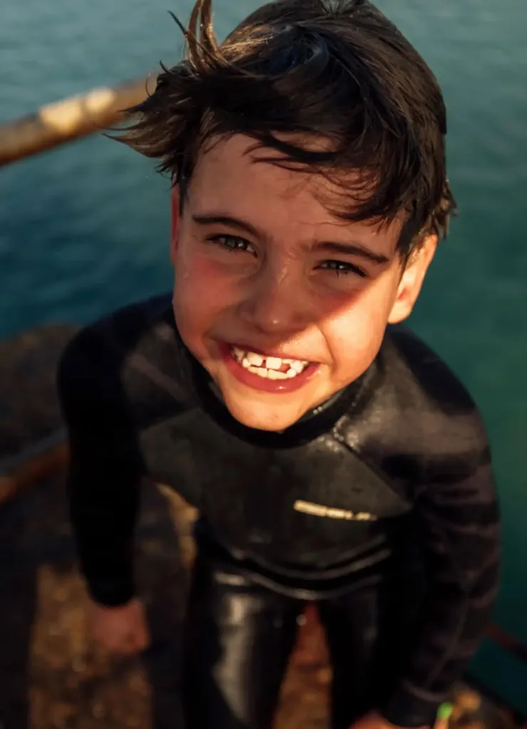 Prince Louis smiling and wearing a wetsuit by the water.