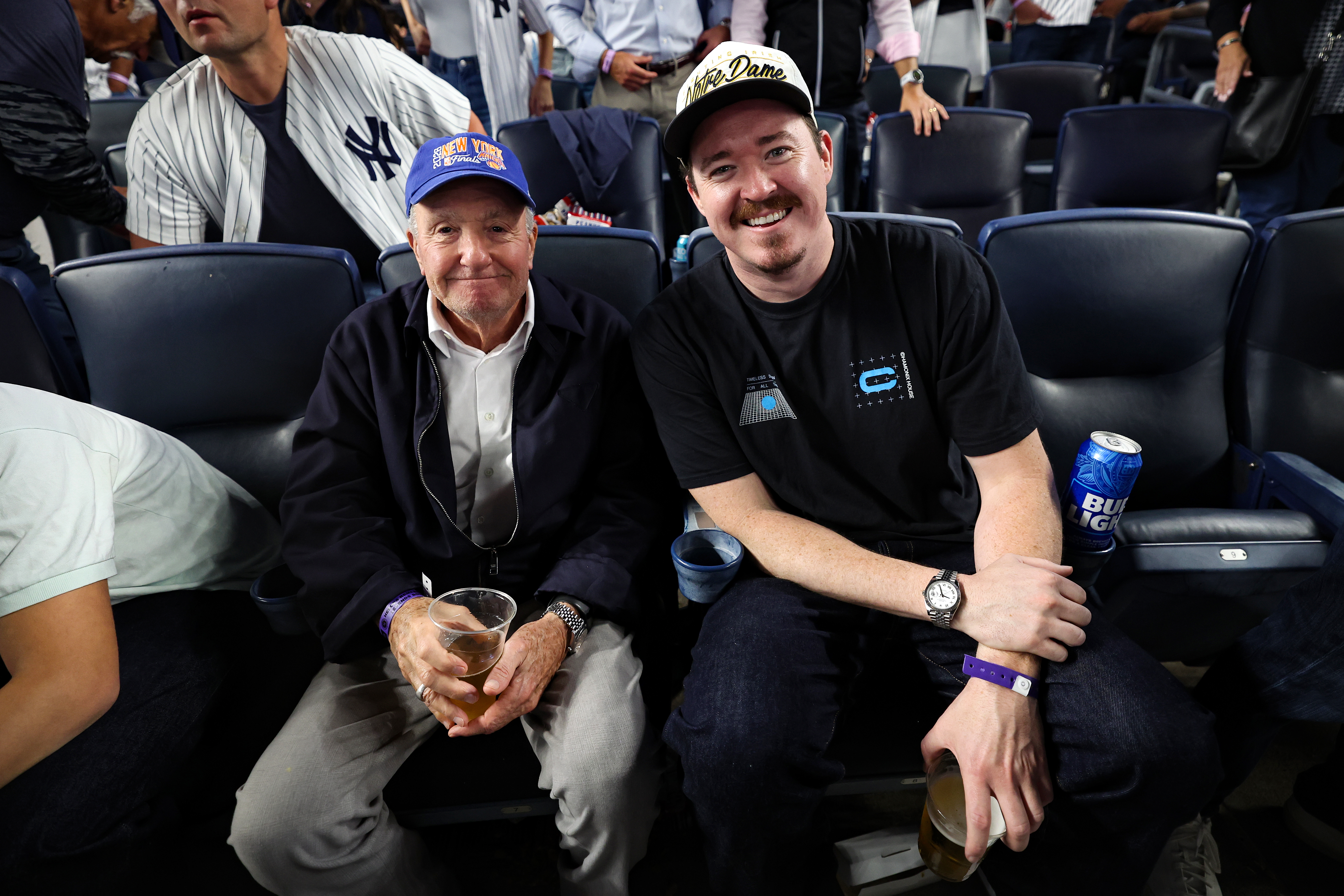 Producer Lorne Michaels and comedian Shane Gillis sit together in stadium seats, holding drinks.