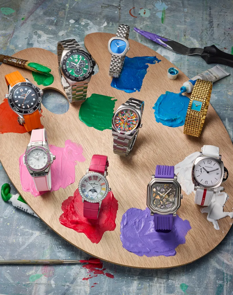 A wooden artist's palette with nine luxury watches arranged on top of colorful dabs of paint, alongside tubes of paint and paintbrushes.