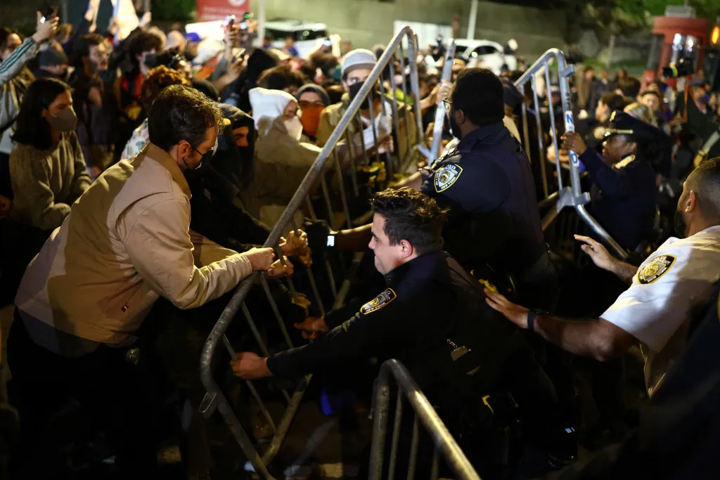 Protesters and law enforcement officers clash across metal barricades in a street at night.