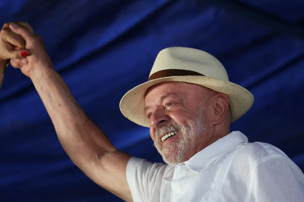 Brazilian President Luiz Inacio Lula da Silva smiles and raises his fist in celebration wearing a light-colored hat and shirt against a blue background.