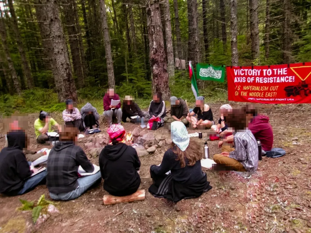 High-school-aged attendees sit in a circle around a campfire in the woods during an 
