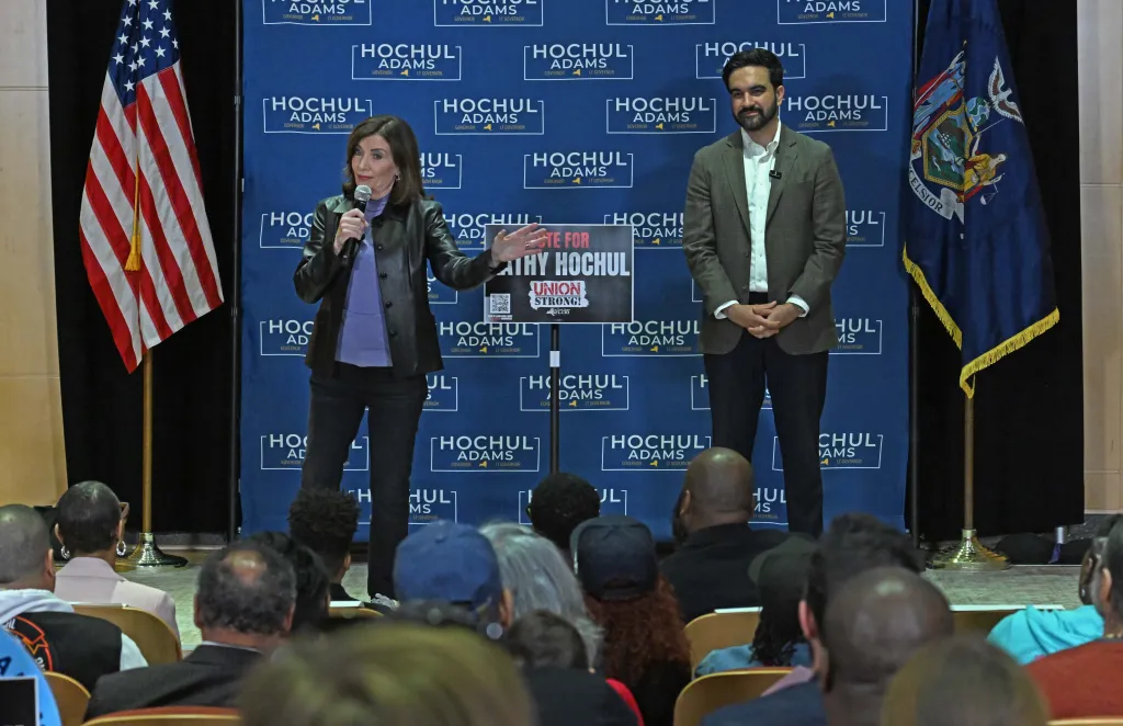 Gov. Hochul speaks into a microphone while Mayor Mamdani stands next to her in front of a crowd.