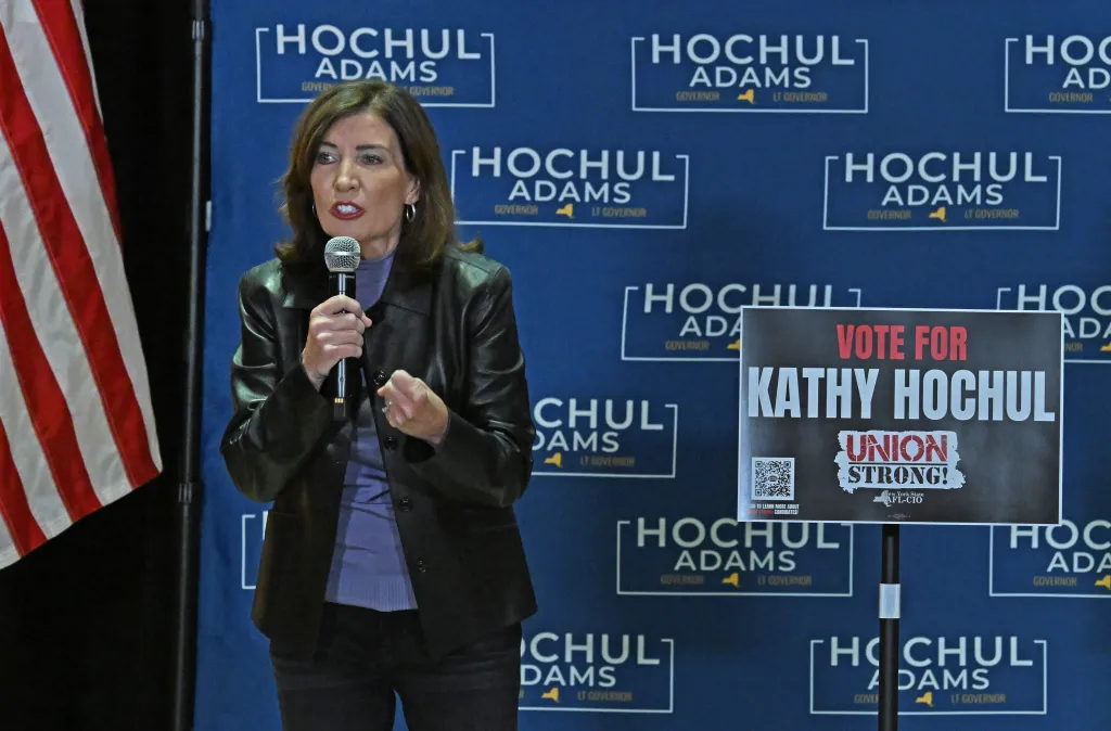 Gov. Kathy Hochul speaks into a microphone at a campaign event, standing in front of a blue backdrop with 