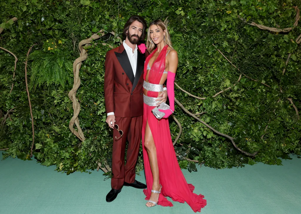 Leonardo Del Vecchio and Jessica Serfaty pose in front of a leafy green backdrop, with Del Vecchio in a maroon suit and Serfaty in a vibrant pink gown with silver accents.
