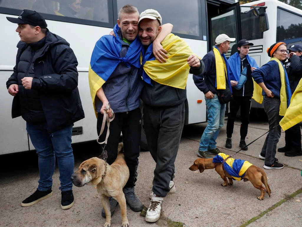 Two evacuated men wrapped in Ukrainian flags embrace outside a bus, accompanied by dogs.