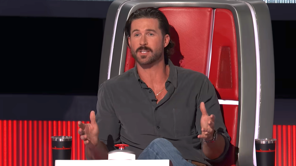 Riley Green sitting in The Voice coaching chair, talking and gesturing with his hands.