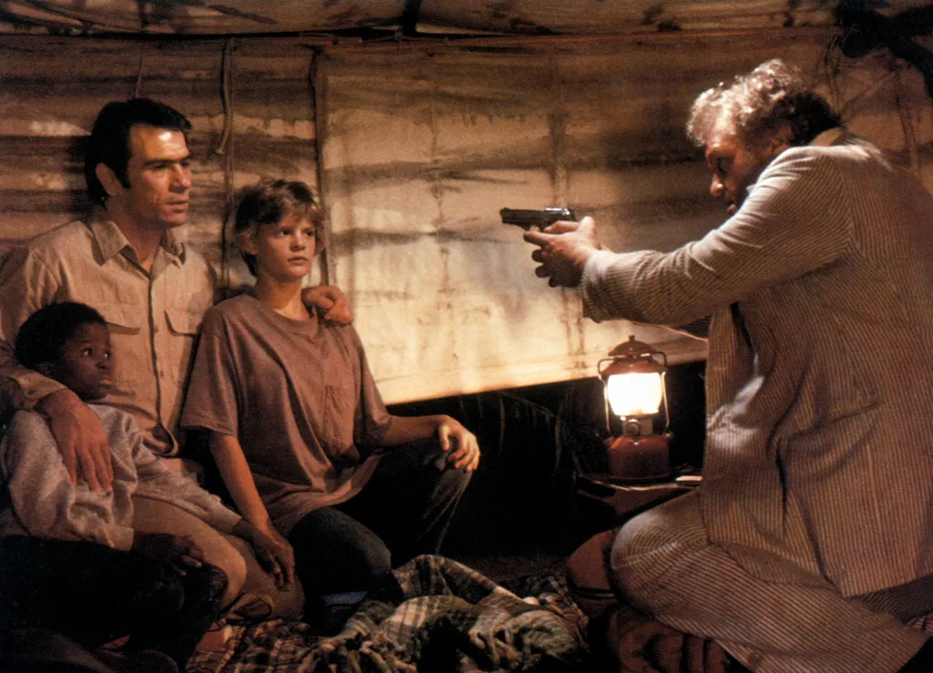 A man points a gun at Tommy Lee Jones, Martha Plimpton, Shawn Smith, and Brian Dennehy inside a tent.