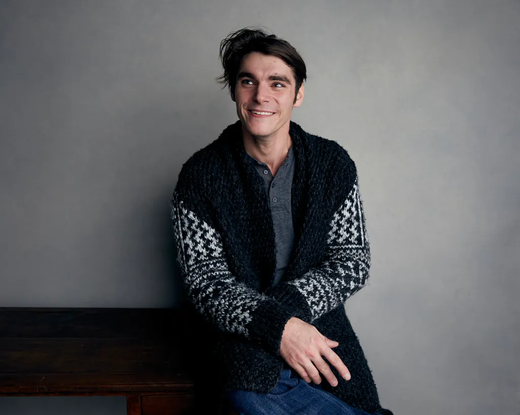 RJ Mitte wearing a patterned cardigan and smiling while looking to the side.