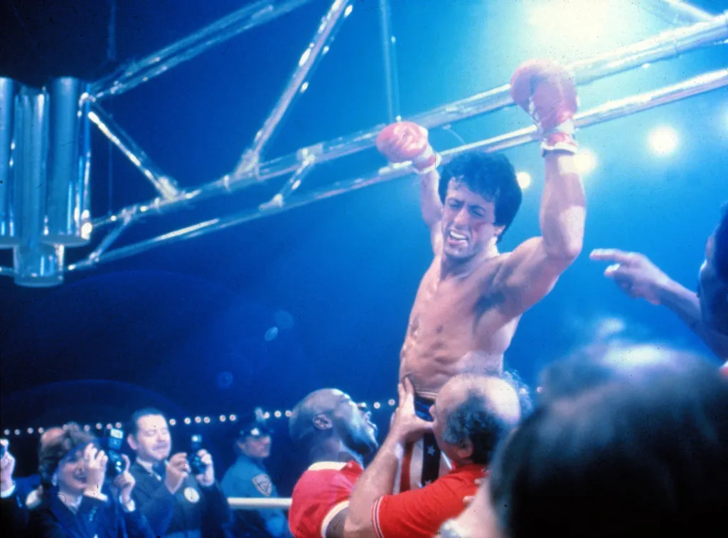 Sylvester Stallone as Rocky Balboa with his arms raised in victory, being carried by his team in the ring.