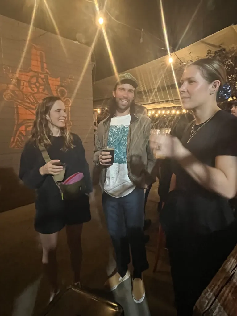 Rosie Bates stands on the left holding a drink, smiling at a man in a cap, while a woman in a black shirt stands to the right, all under string lights at night.