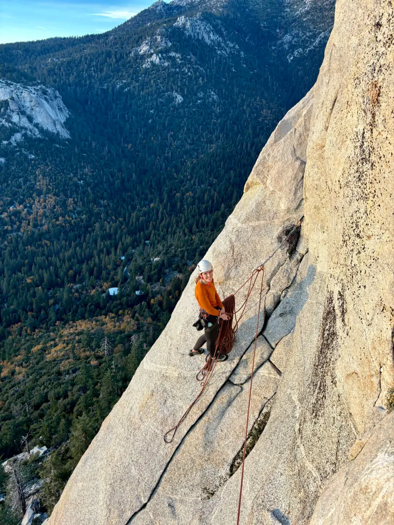 Rosie Bates poses on a steep granite cliff face with climbing gear, overlooking a vast pine forest valley.