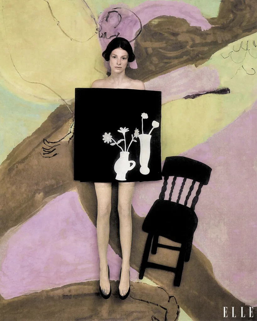 Hunter Schafer wearing a black dress featuring a white still life graphic against a painted backdrop, posing next to a black chair.