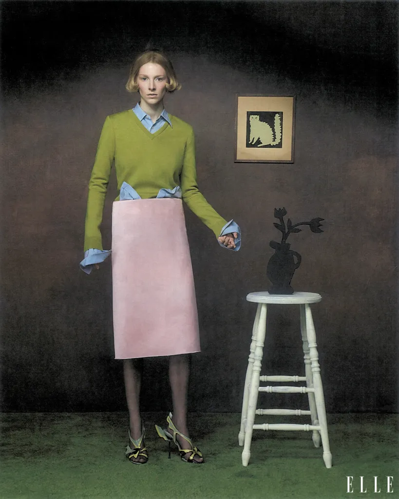 Hunter Schafer poses for ELLE, wearing a green sweater over a blue shirt and a pink skirt, standing beside a stool with a vase.