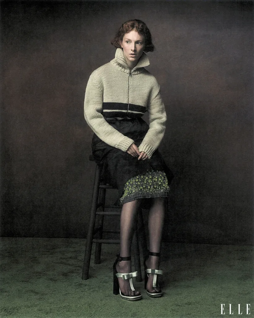 Hunter Schafer wearing a cream-colored zip-up sweater and dark skirt, seated on a wooden stool against a dark backdrop.