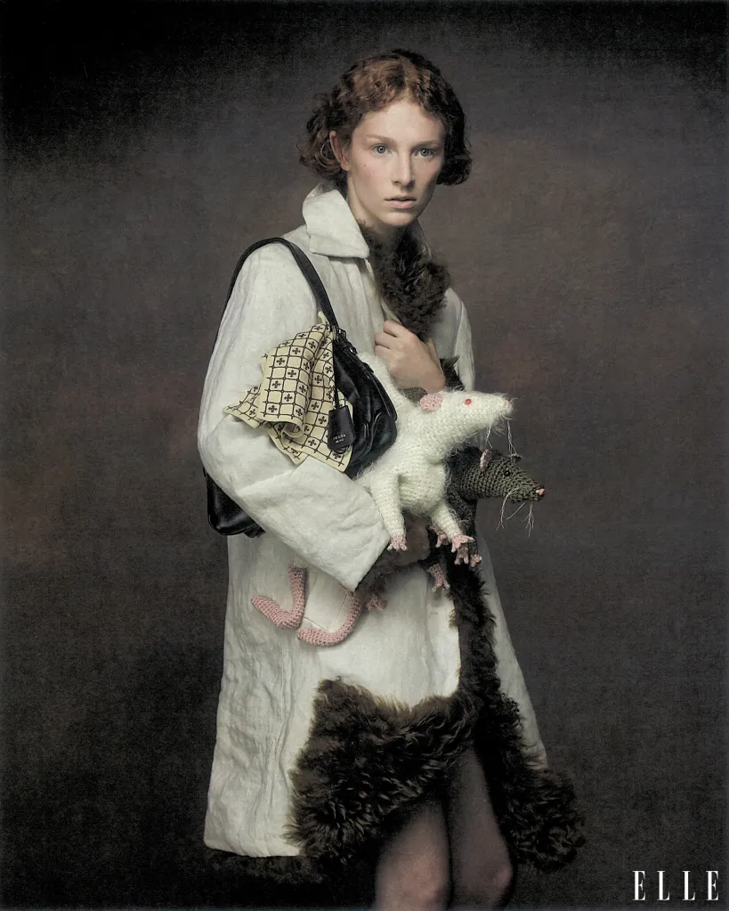Hunter Schafer wearing a white coat with fur trim and holding two knitted rats.