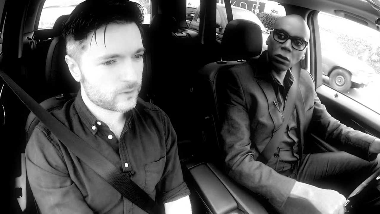 Lucian Piane and RuPaul sitting in a car in a black-and-white still image.