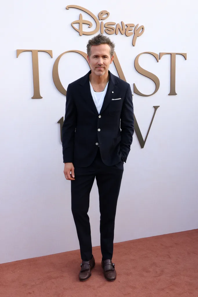 Ryan Reynolds poses in a dark suit over a white t-shirt in front of a Disney Toast to TV backdrop.