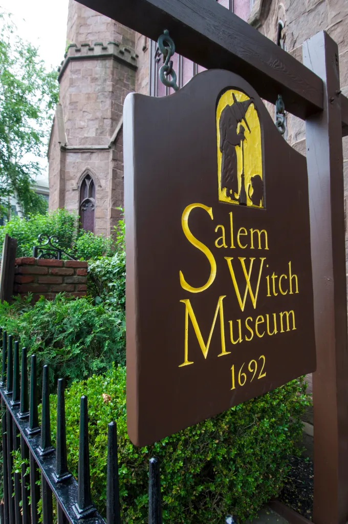 A wooden sign for the Salem Witch Museum featuring a yellow silhouette of a witch with a broomstick, with the museum's stone building and lush greenery in the background.