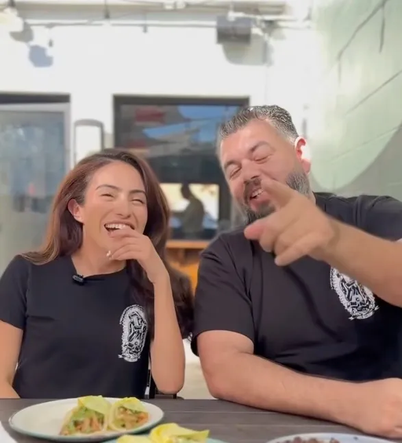Sasha Renée Pérez and a man wearing LA Taco shirts laughing and eating tacos at an outdoor table.
